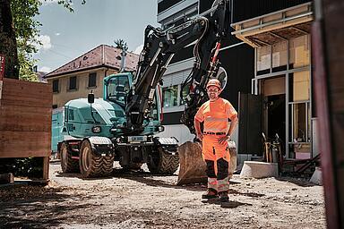 Ein Maschinenführer in Warnschutzkleidung steht neben einem Mobilbagger vom Typ Wacker Neuson EW100 auf einer städtischen Baustelle.