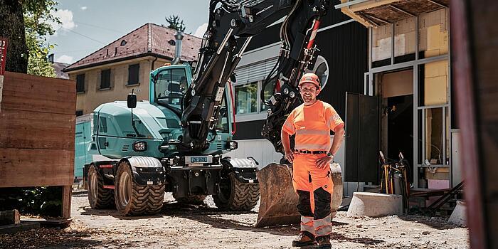 Ein Maschinenführer in Warnschutzkleidung steht neben einem Mobilbagger vom Typ Wacker Neuson EW100 auf einer städtischen Baustelle.