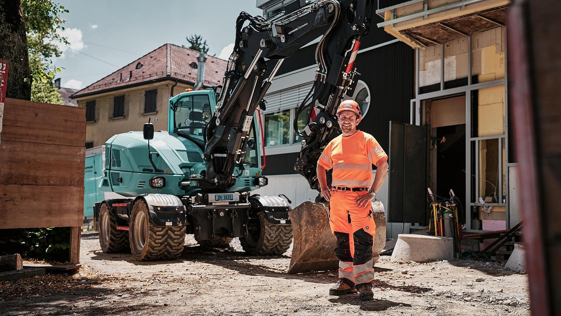 Ein Maschinenführer in Warnschutzkleidung steht neben einem Mobilbagger vom Typ Wacker Neuson EW100 auf einer städtischen Baustelle.
