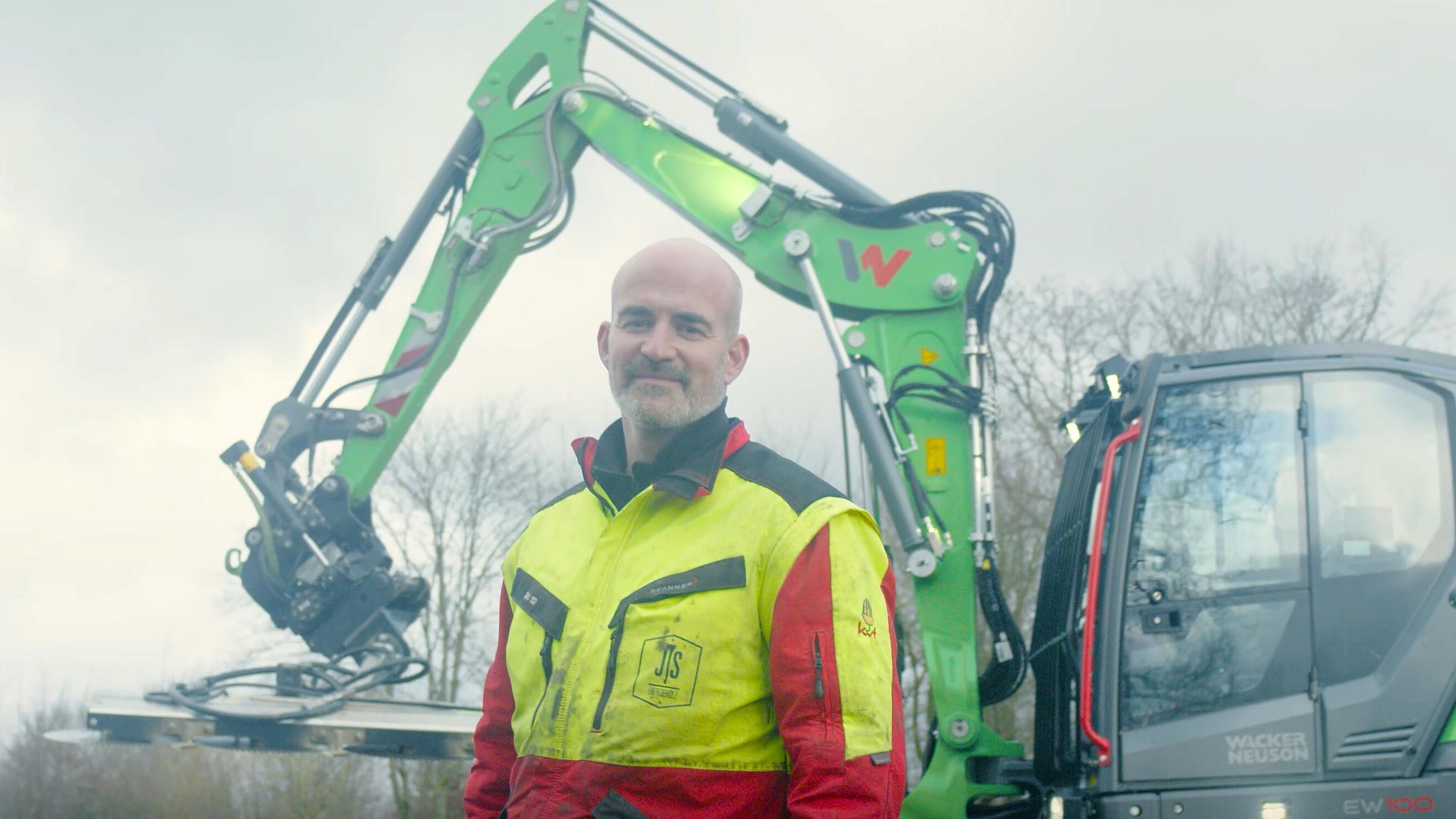 Ein Maschinenführer steht vor einem Mobilbagger vom Typ Wacker Neuson EW100, der mit einem Tiltrotator als Anbaugerät ausgestattet ist.
