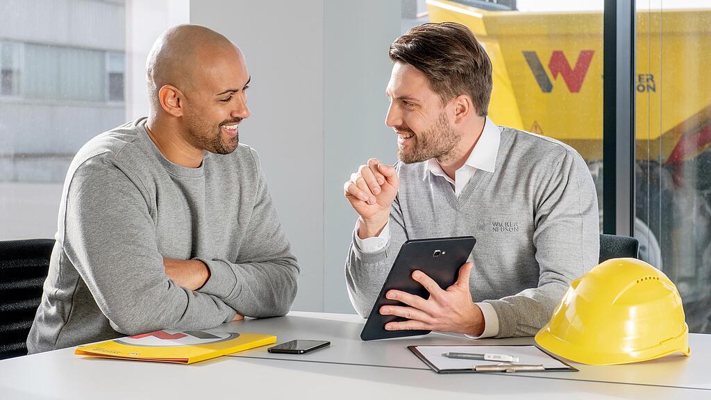 Wacker Neuson Mitarbeiter sitzt am Schreibtisch und zeigt Kunden etwas auf dem Tablet.