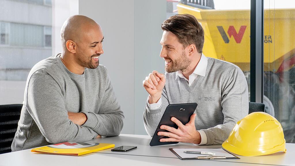 Wacker Neuson Mitarbeiter sitzt am Schreibtisch und zeigt Kunden etwas auf dem Tablet.