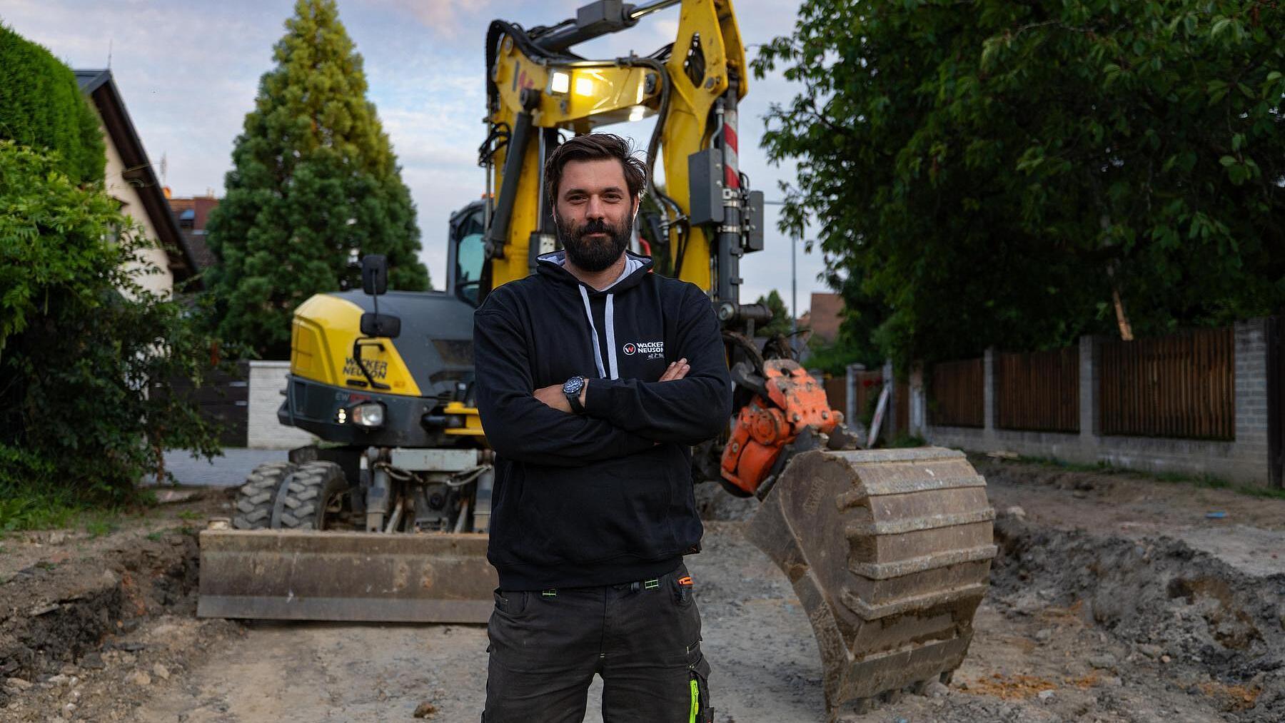 Ein Baufachmann steht auf einer Baustelle vor einem Mobilbagger vom Typ EW100 von Wacker Neuson.